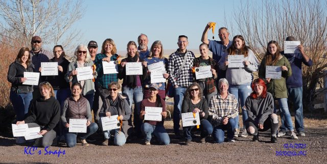 About two dozen owners outside at the ranch on a sunny winter day, holding up their ZASSC certificates; several hold medallions on orange ribbons.